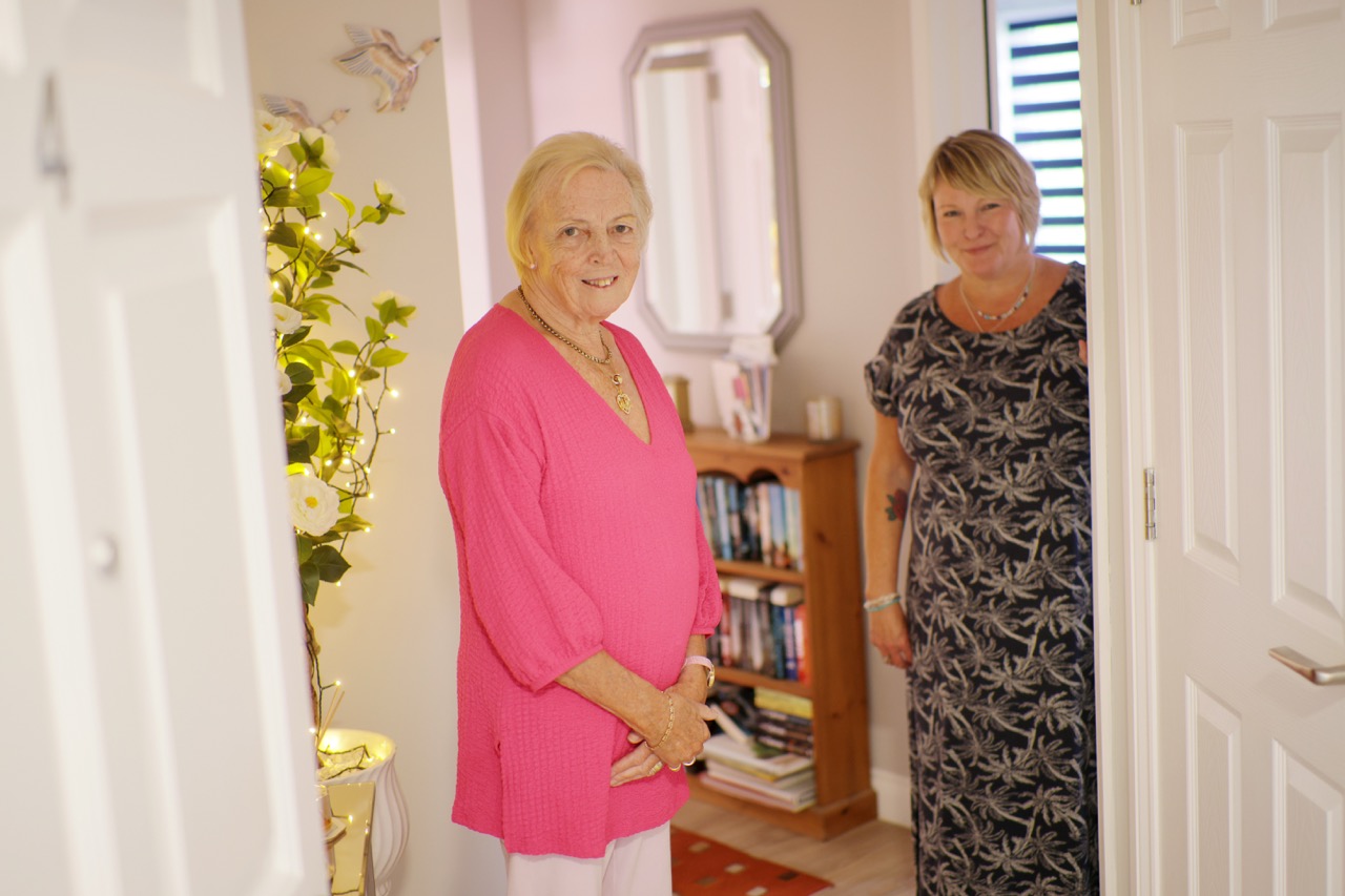 Two smiling women indoors, one in a pink top standing near a plant with fairy lights, the other in a patterned dress standing by a door.