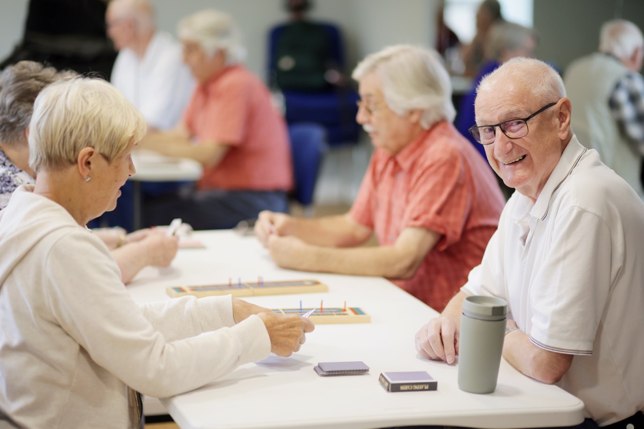Elderly people sitting at tables playing card games in a community center.