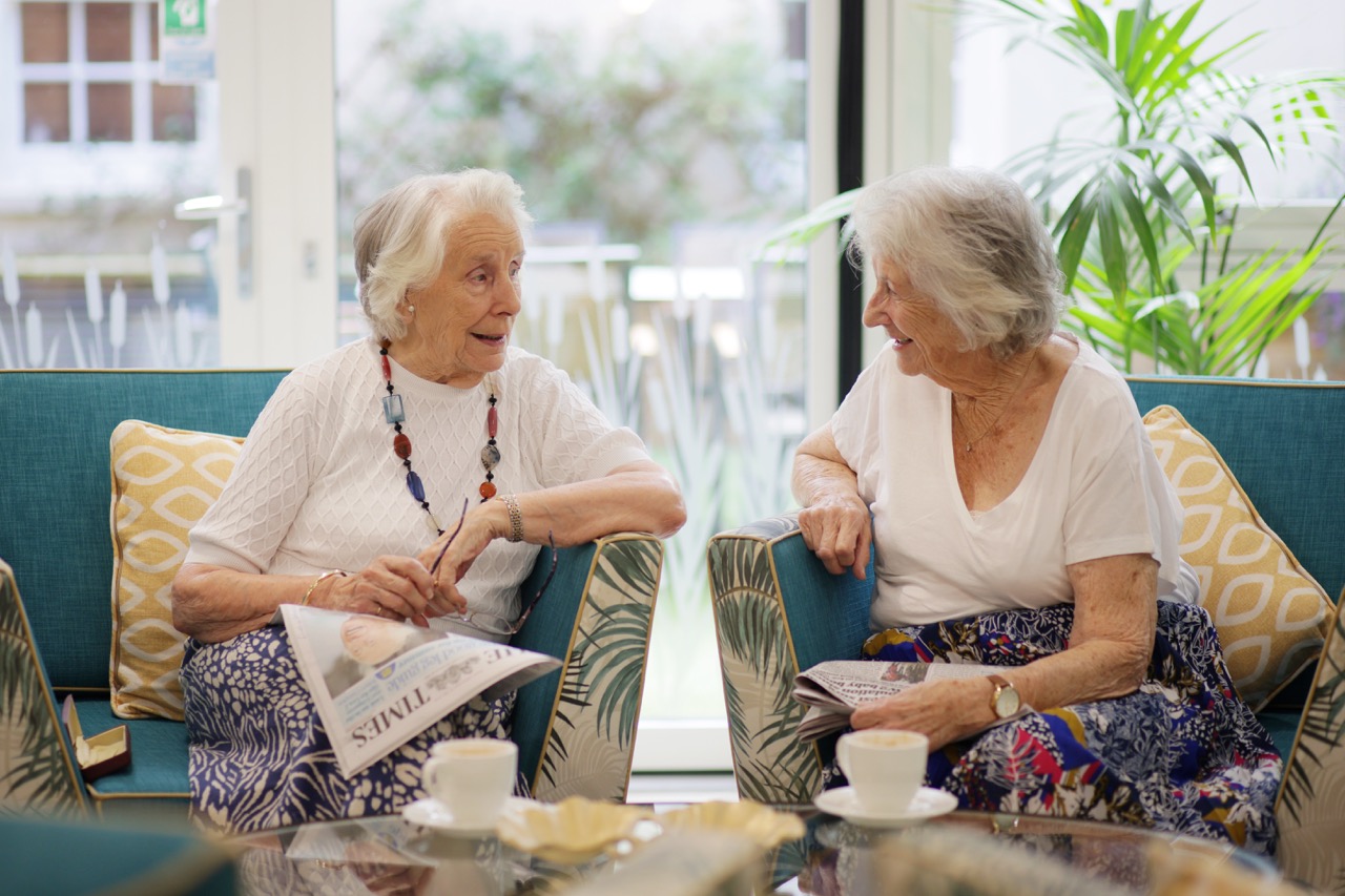 Two elderly women sitting on blue armchairs in a bright room, smiling and talking while holding newspapers with coffee cups on a glass table in front of them.