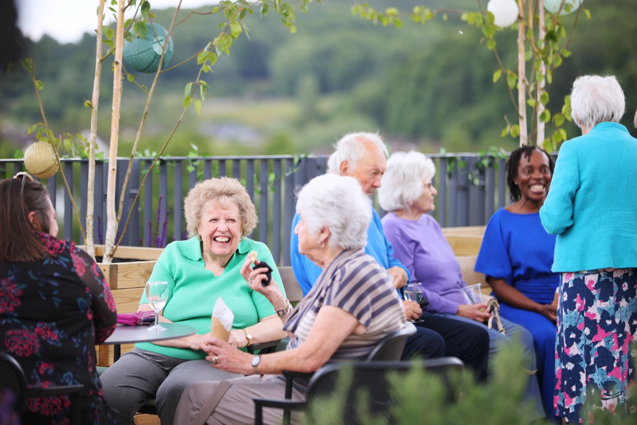 Smiling elderly man with glasses sitting on a blue armchair reading a newspaper indoors.
