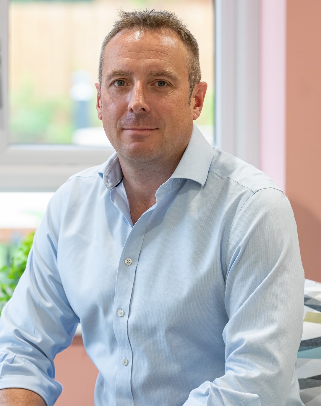 Man in a light blue shirt sitting indoors near a window with a neutral expression.