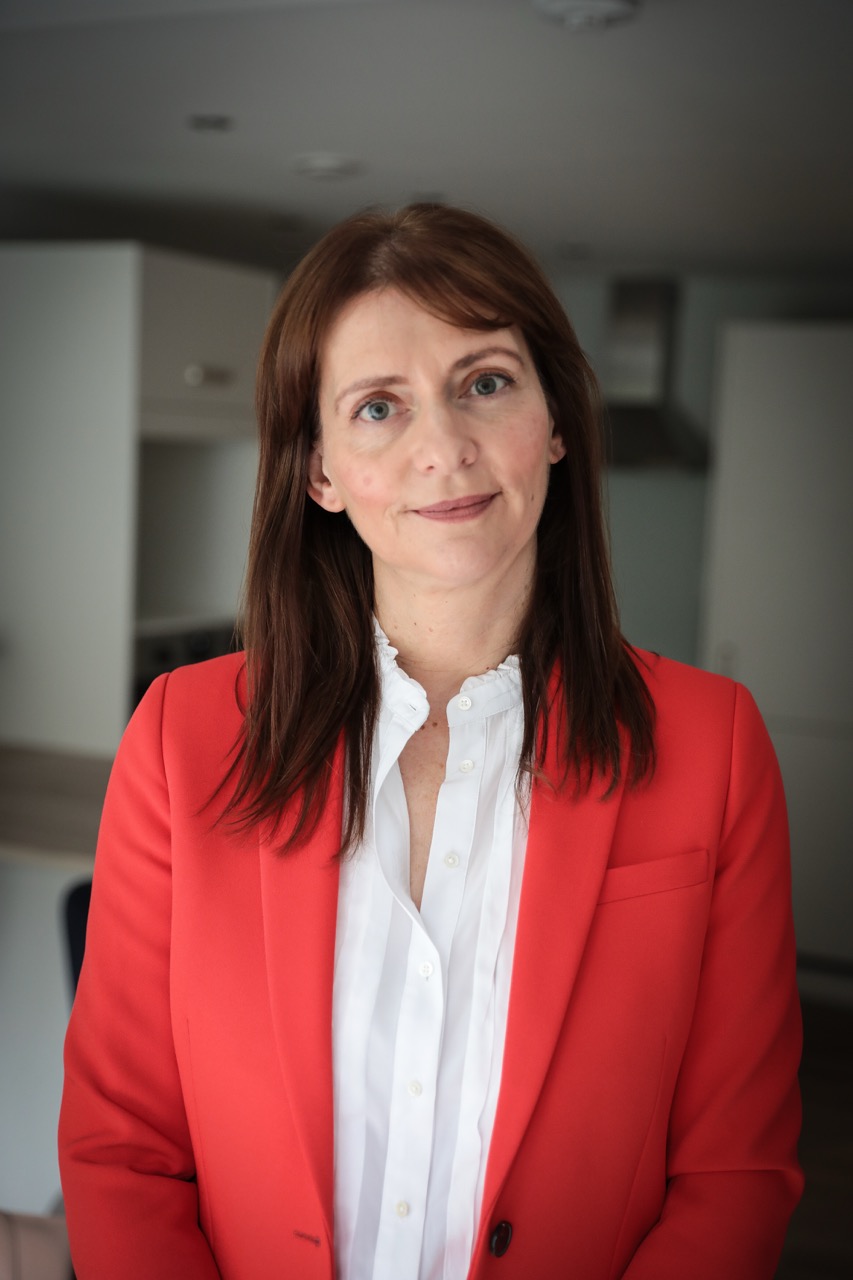 Smiling woman with long brown hair wearing a red blazer and white shirt standing indoors.