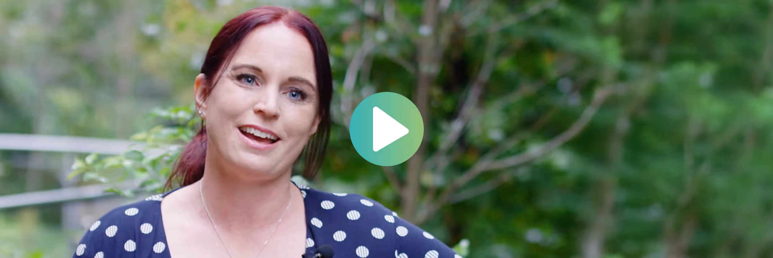 Smiling woman with red hair tied back standing outdoors in a green, leafy background.