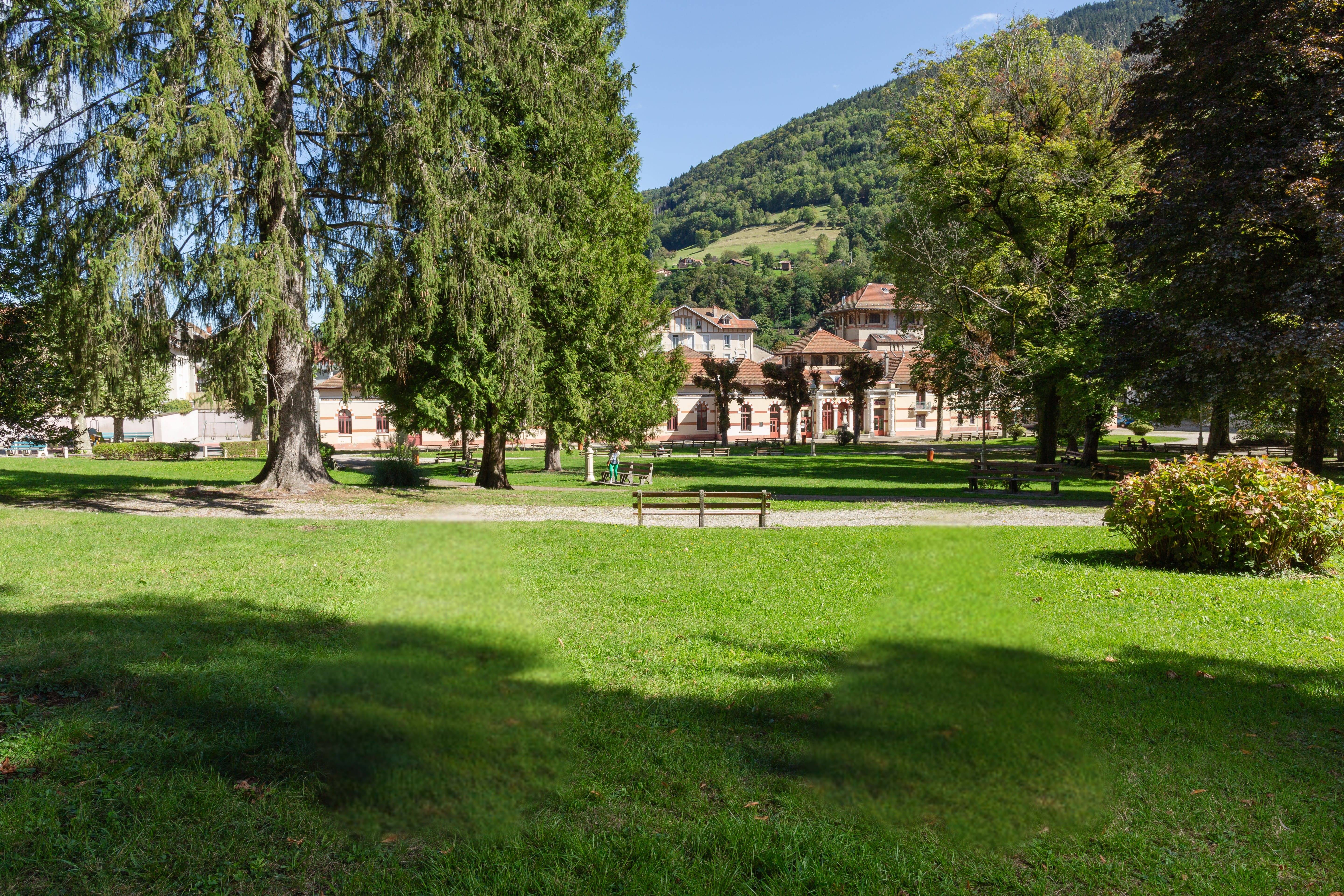 Parc verdoyant avec des bancs, de grands arbres et un bâtiment traditionnel au pied d'une colline boisée sous un ciel bleu.