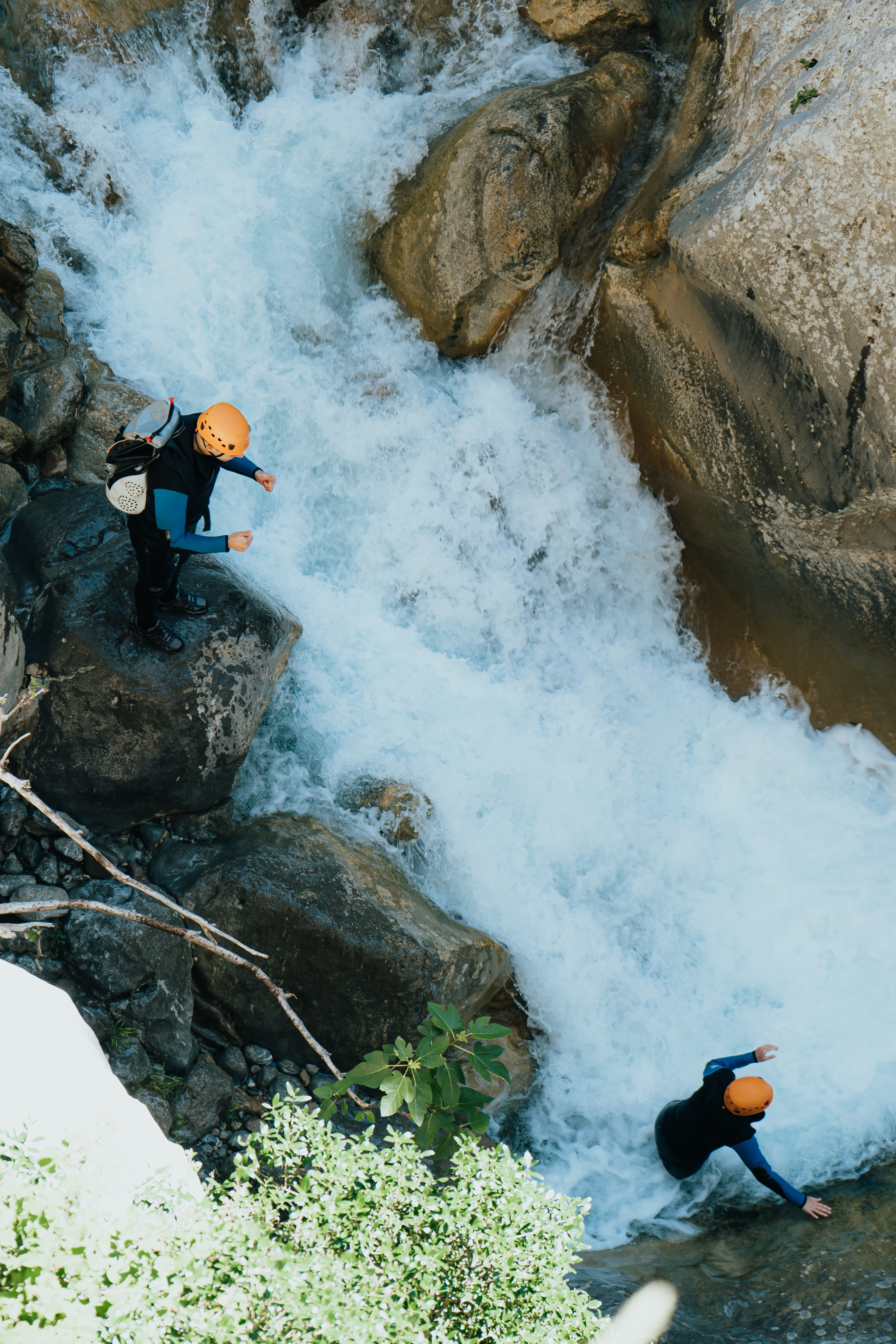 Deux personnes portant des casques orange pratiquent le canyoning dans une rivière tourbillonnante avec des rochers autour.