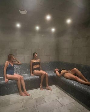 Trois femmes en maillots de bain assises et allongées dans un hammam aux murs et bancs carrelés, avec de la vapeur légère.