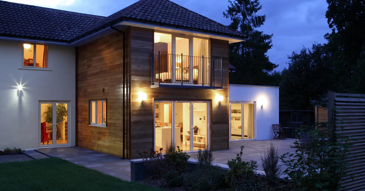 Grande maison contemporaine éclairée à la tombée du jour : bardage bois, larges baies vitrées, terrasse en pierre et jardin arboré
