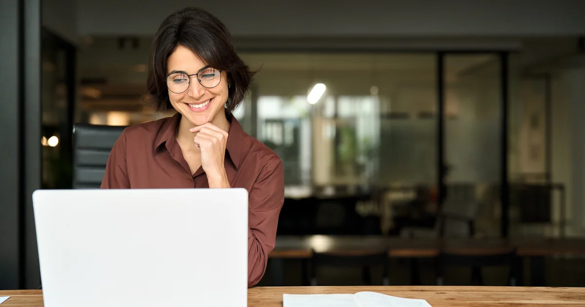 Professionnelle de l'immobilier souriante devant un ordinateur portable, dans un bureau aux cloisons vitrées