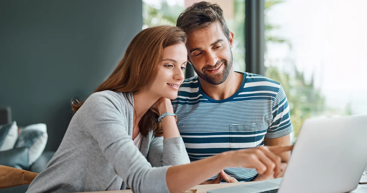 Couple souriant assis devant un ordinateur portable, la femme montre l'écran du doigt, papiers et lunettes posés sur la table, intérieur lumineux