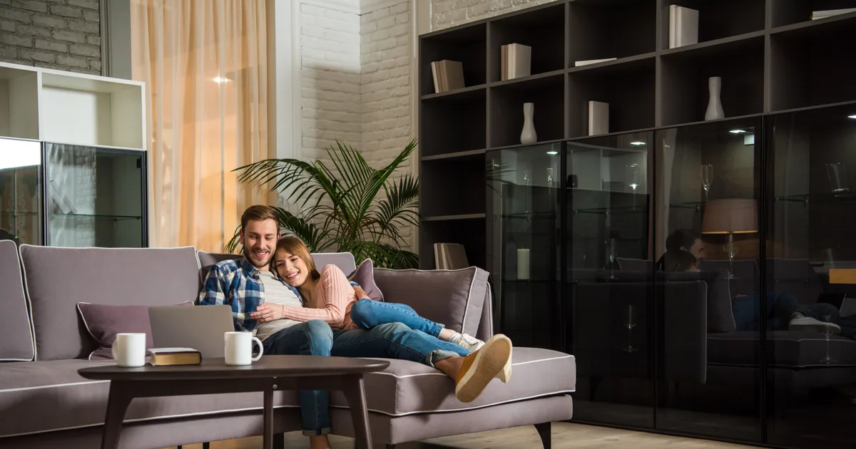 Jeune couple souriant installé sur un canapé, un ordinateur portable ouvert sur la table basse, dans un salon aux grandes étagères