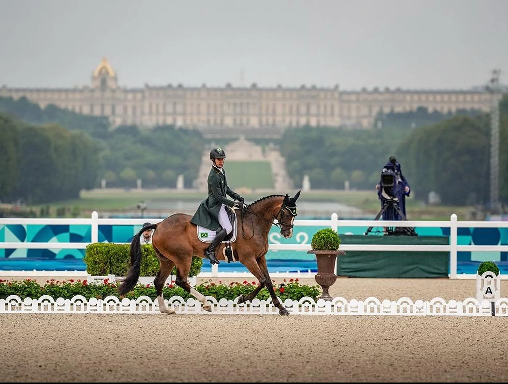 Equestrian rider in formal attire performing dressage on a brown horse in an outdoor arena with a historic palace and garden in the background.