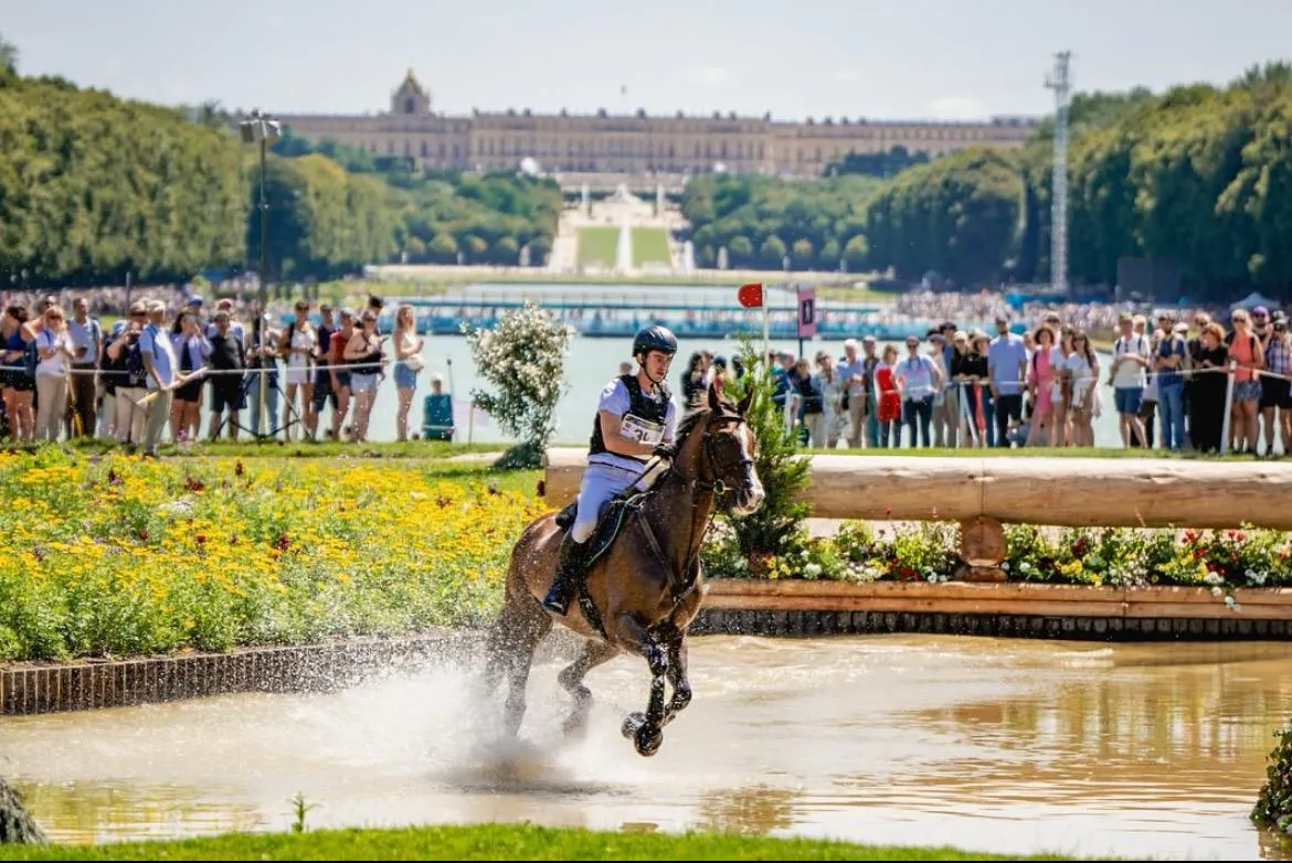 Horse and rider jumping through a water obstacle in an equestrian event with a large crowd and historic building in the background.