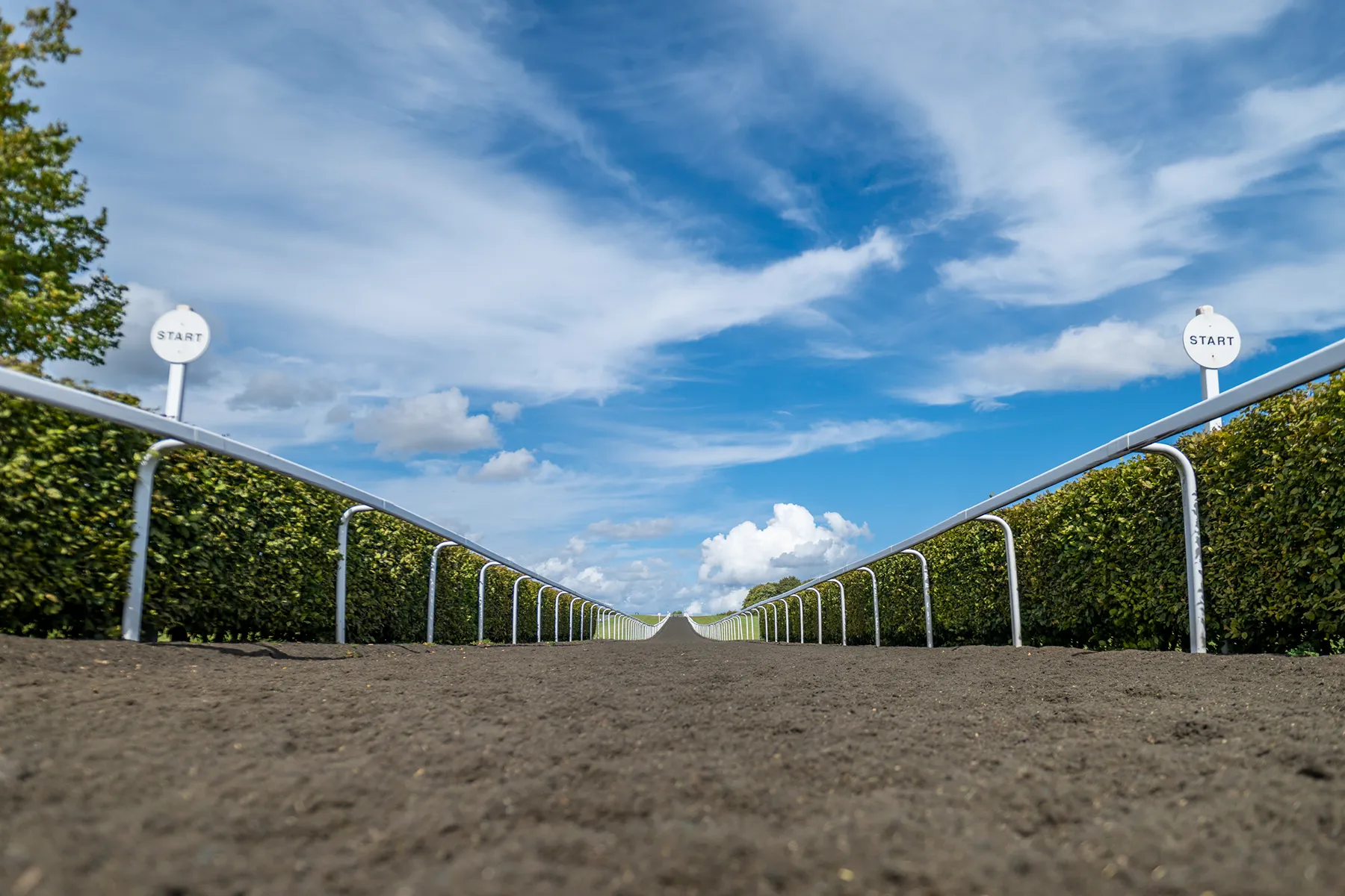 Low-angle view of a dirt racetrack with metal railings and two 'START' signs against a blue sky with clouds.