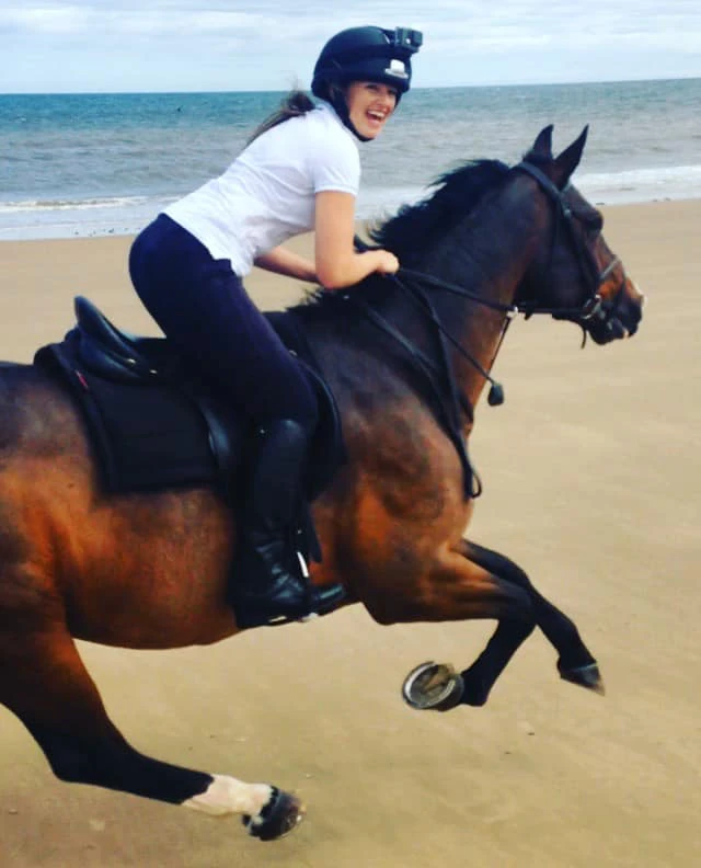 Smiling person wearing a helmet riding a galloping brown horse on a sandy beach near the ocean.