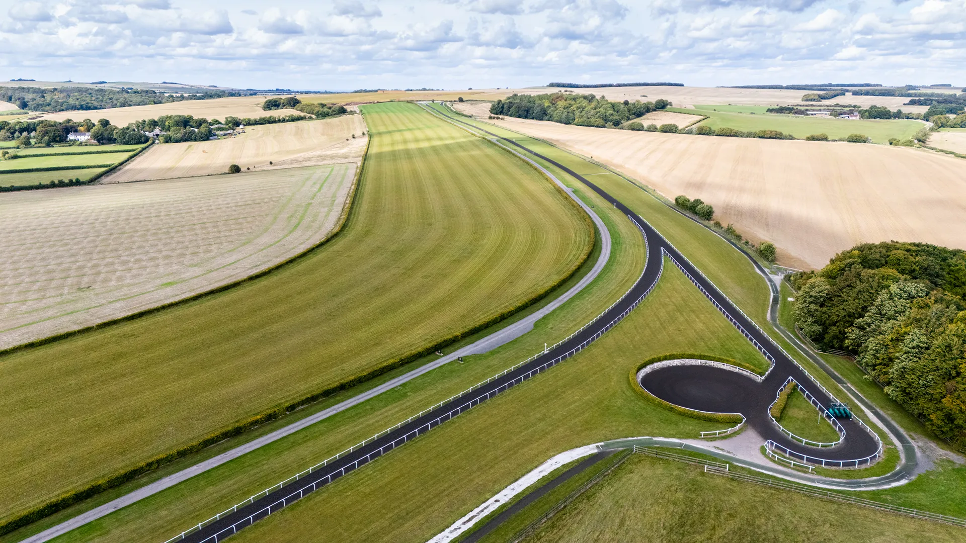 Aerial view of a winding racetrack surrounded by green and golden fields under a partly cloudy sky.