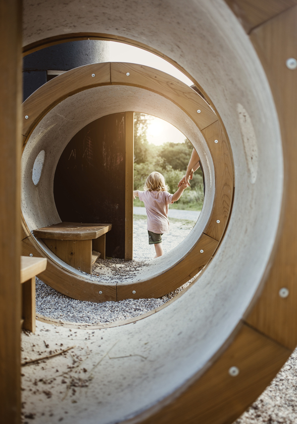 Bambino che cammina tenendo la mano di un adulto attraverso grandi tubi di cemento in un parco giochi all'aperto al tramonto.