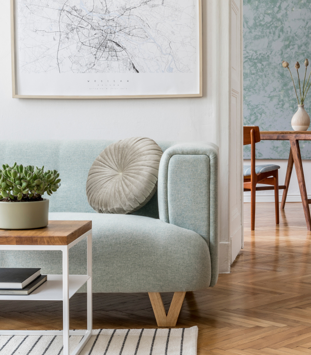 Divano turchese con cuscino rotondo beige, tavolino in legno e metallo con pianta succulenta e libri, parete bianca con mappa incorniciata, pavimento in parquet e vista di una stanza adiacente con tavolo e sedie.