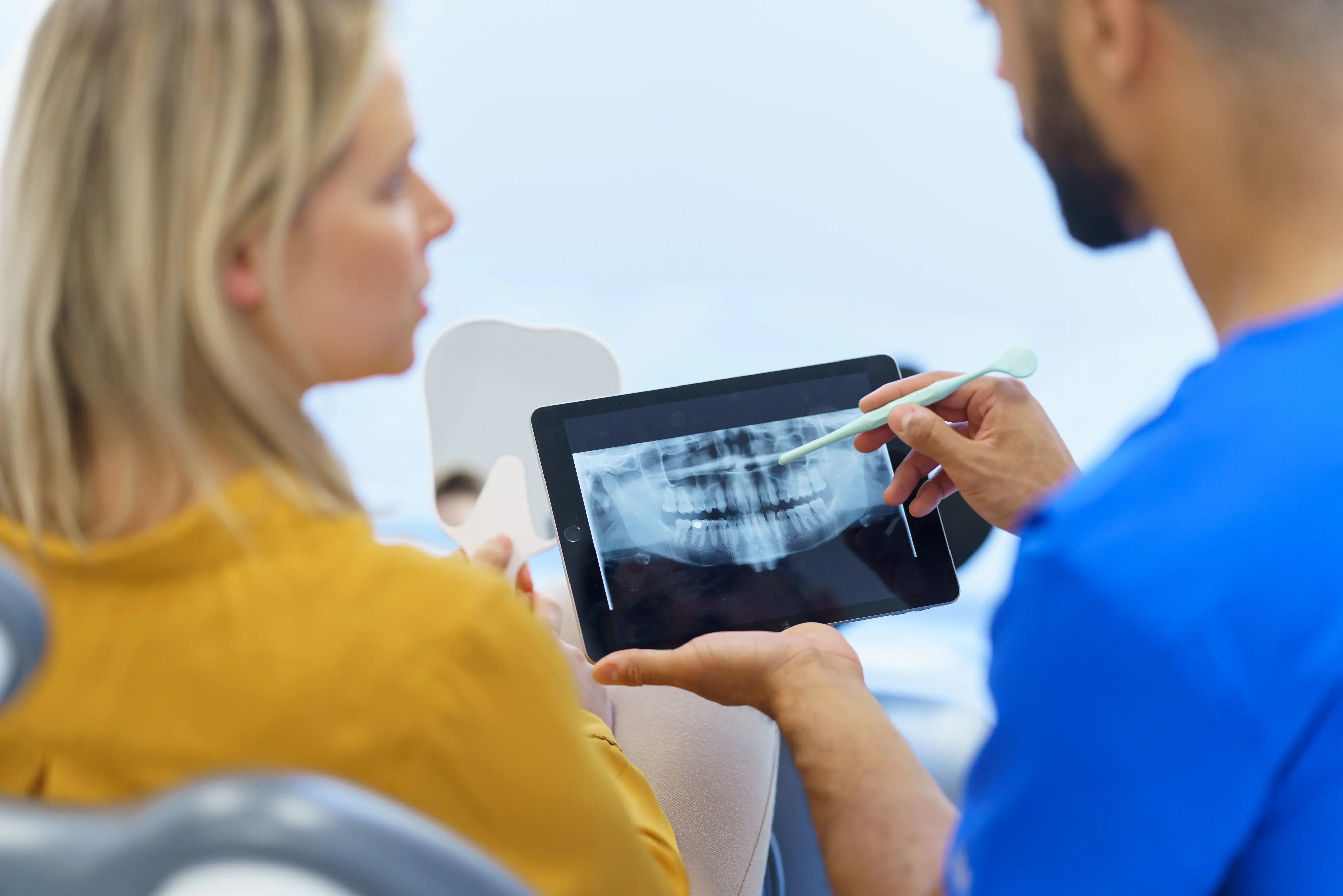 A patient looks at a digital X-ray on a tablet while the dentist explains the panoramic scan, highlighting areas of concern in the teeth and jaw.