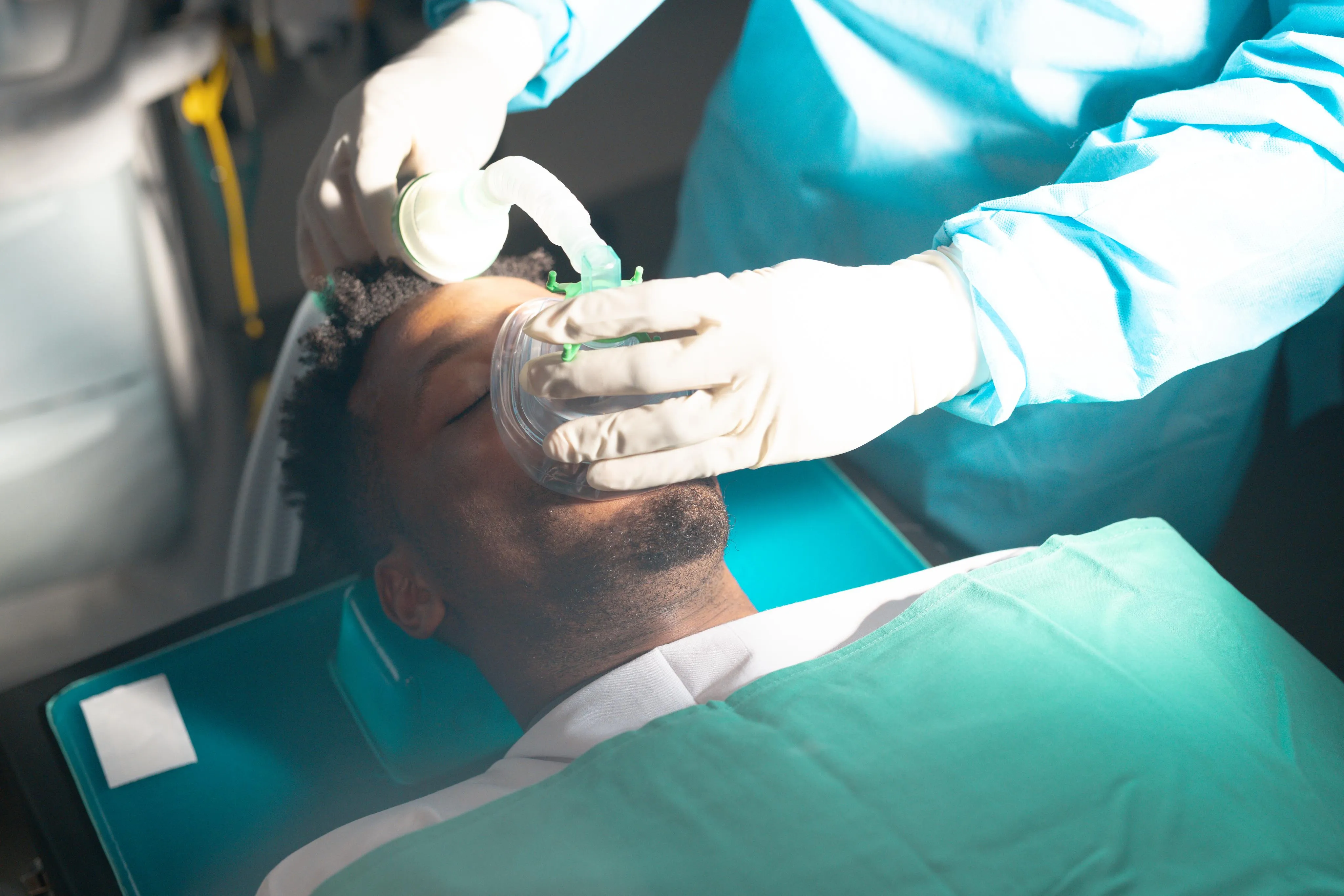 A patient lying on a dental chair receives sedation as a clinician places an oxygen and anesthesia mask over his nose and mouth, preparing him for a relaxed and comfortable dental procedure.