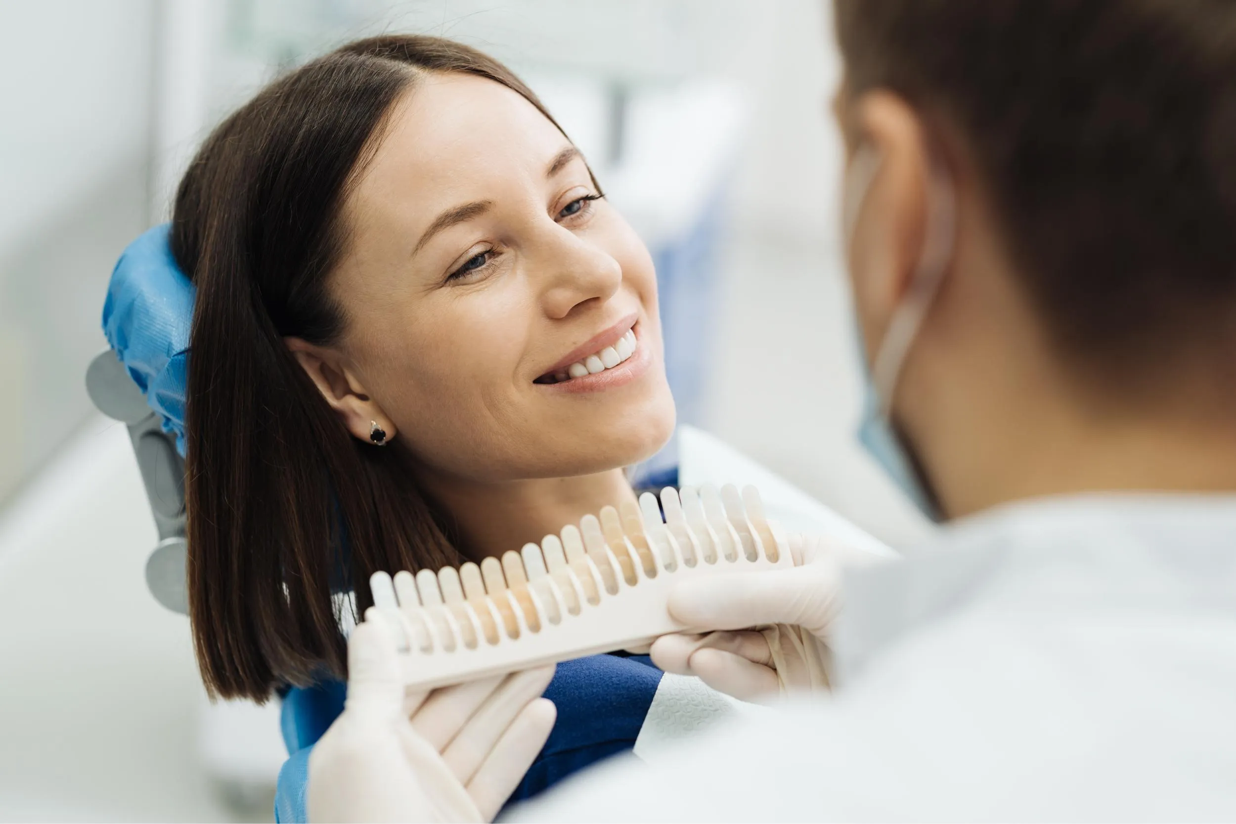 A patient in the dental chair smiles as the clinician holds a veneer shade guide next to her teeth to determine the most natural-looking veneer color.