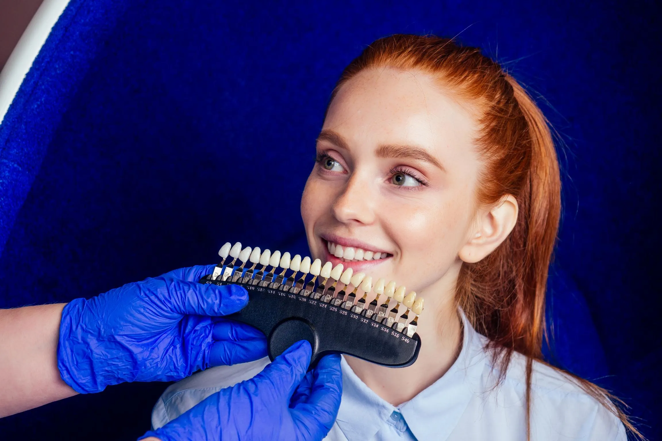 A smiling patient holds up a shade guide while a dental professional compares tooth colors to select the ideal shade for porcelain veneers.