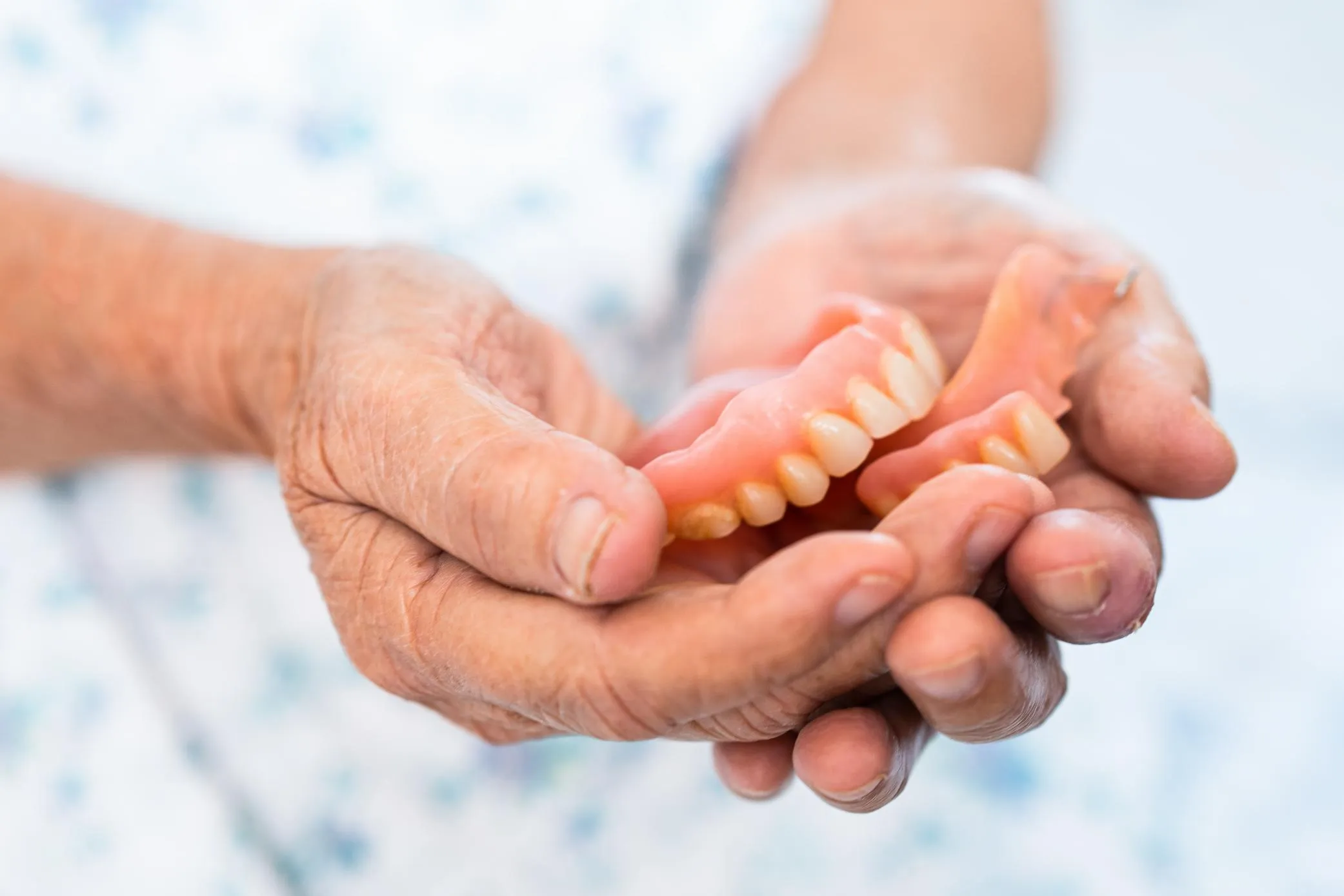 An older adult holding a set of full dentures in their hands, highlighting the realistic tooth design and gum-colored base of removable dentures used to replace missing teeth.