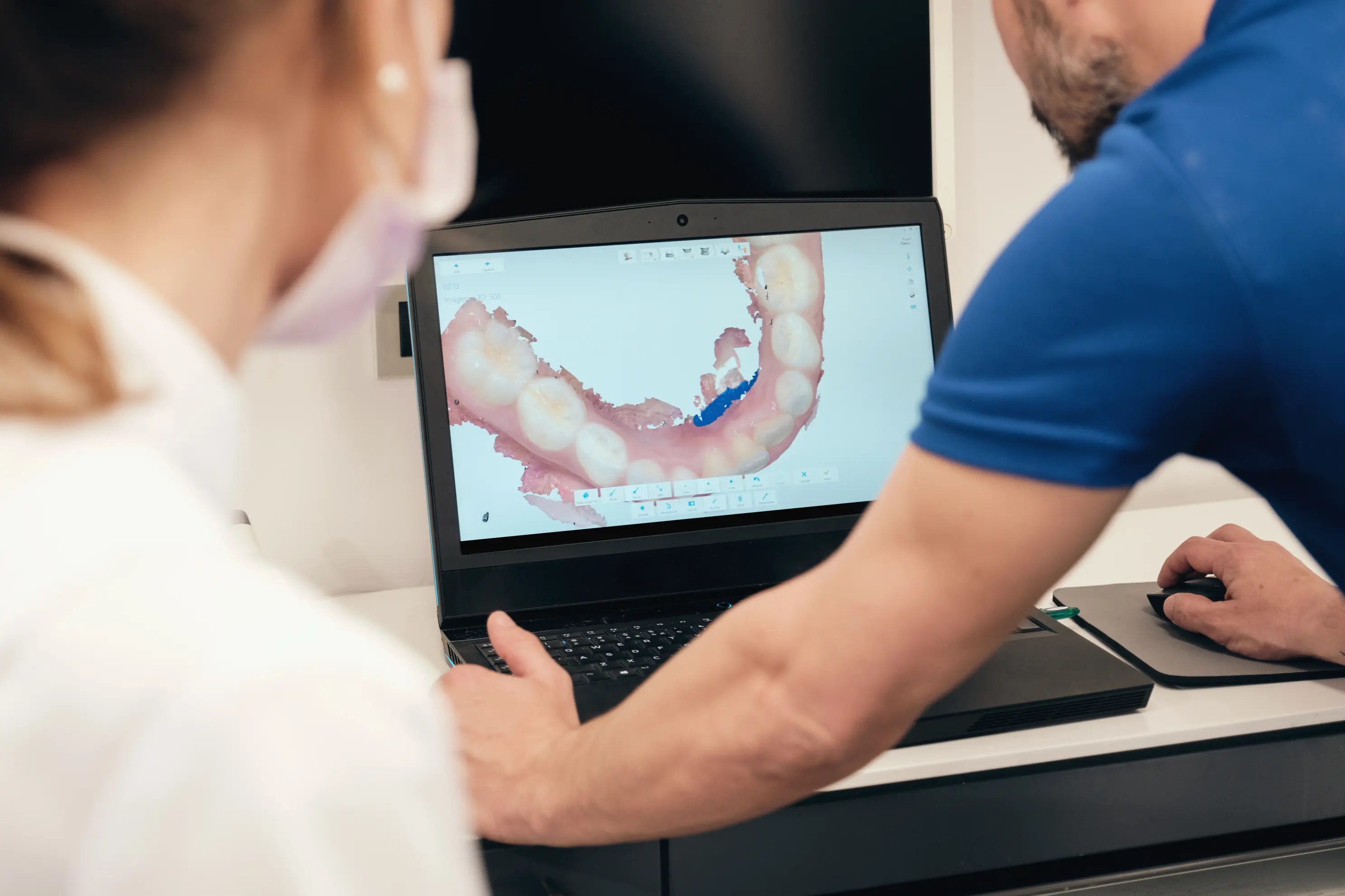 A dentist and patient looking at a laptop screen showing a full-color 3D model of the patient’s jaw, demonstrating how computer-guided implant technology maps ideal implant angles and placement sites.