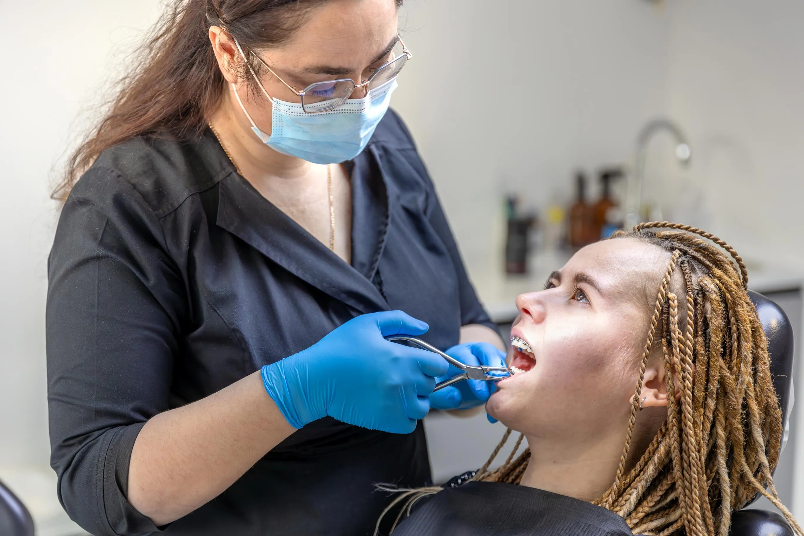 An orthodontic clinician wearing a mask and gloves examines a patient’s teeth while adjusting their metal braces during a routine orthodontic appointment.