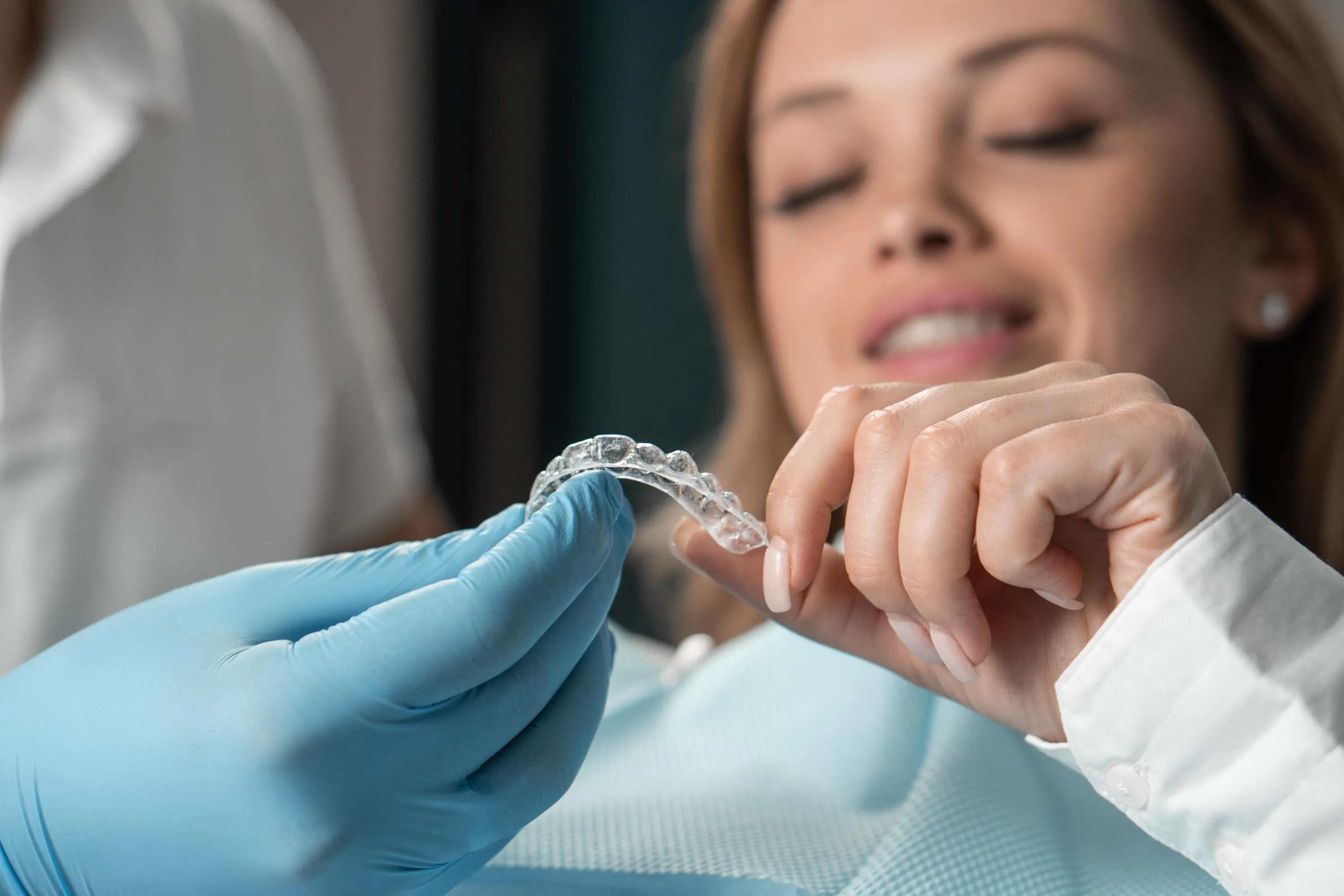 A patient holds a transparent clear aligner tray while a clinician in blue gloves guides her through how to insert and remove the aligner, showing the flexibility and custom fit of orthodontic clear aligners.