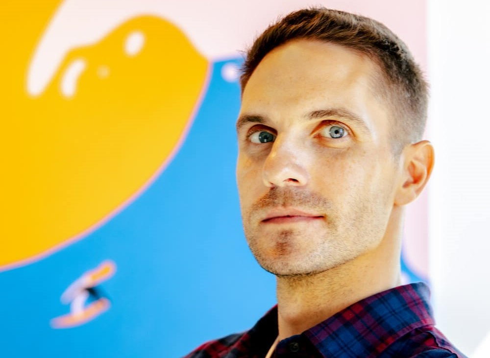 Close-up portrait of a man standing in front of a colorful abstract mural, looking slightly off camera.
