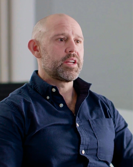 Bald man with a beard wearing a dark blue button-up shirt speaking indoors.