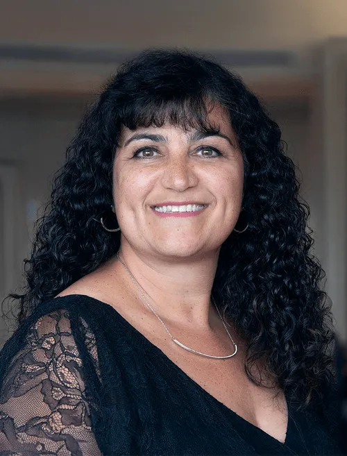 Smiling woman with curly black hair wearing a black lace top and silver necklace in an indoor setting.