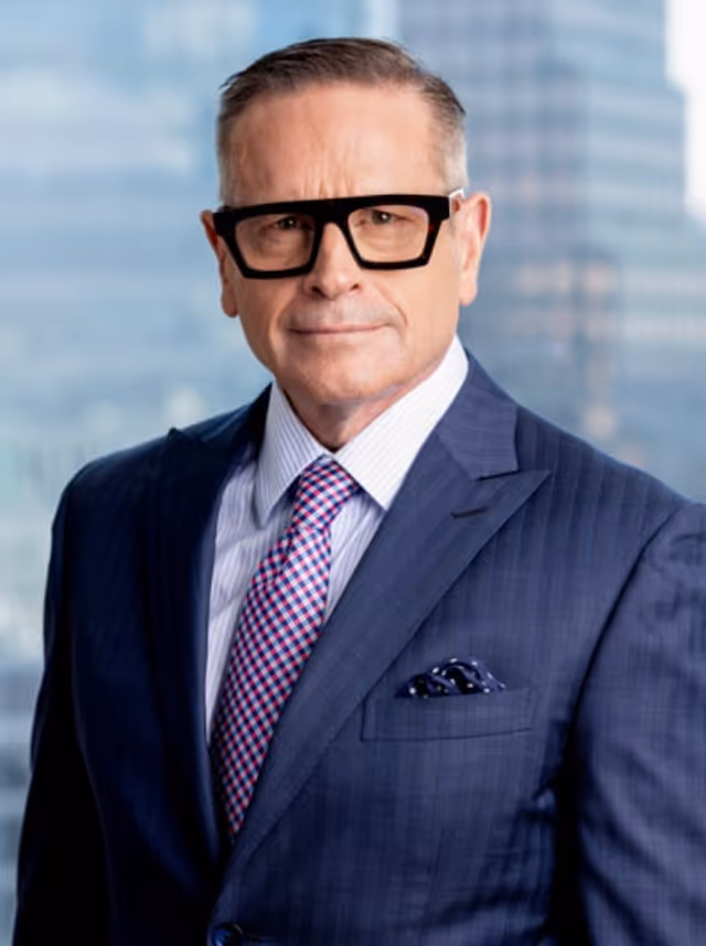 Middle-aged man wearing large black glasses, a navy pinstripe suit, a blue and pink patterned tie, and a white shirt, standing in front of a blurred cityscape background.