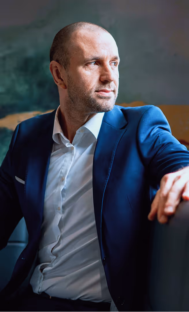Man in a blue suit and white shirt looking thoughtfully to the side while seated indoors.