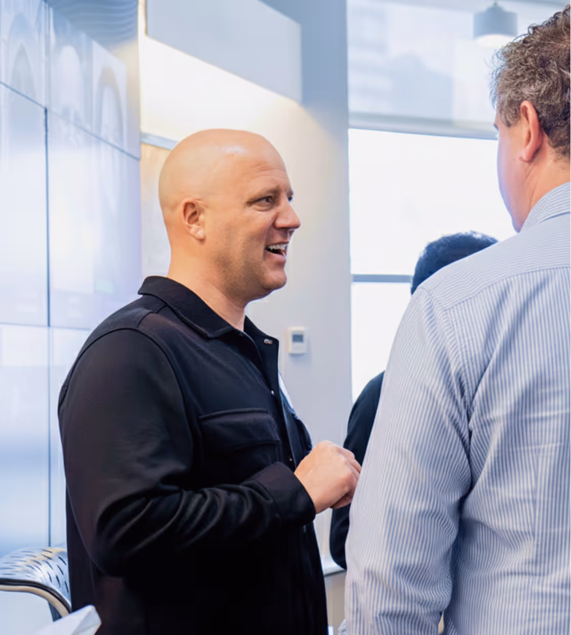 Man in a black shirt smiling and talking to another man in a striped shirt in a bright indoor setting.