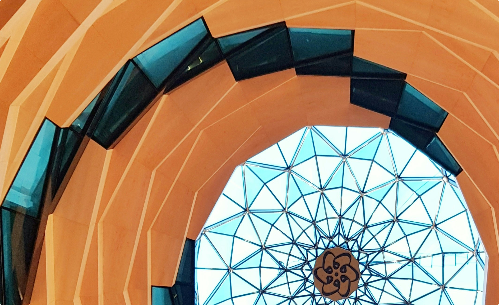 Architectural ceiling featuring an orange geometric wooden frame encircling a glass dome with a metal lattice and central floral design.