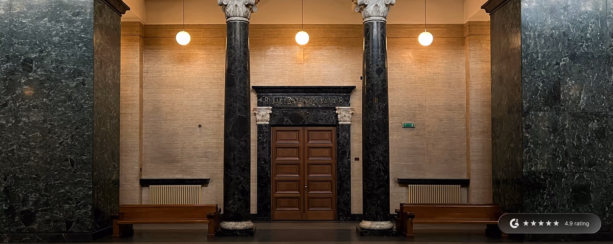 Interior of a courtroom hallway with wooden double doors framed by dark marble columns and beige walls, illuminated by hanging globe lights.