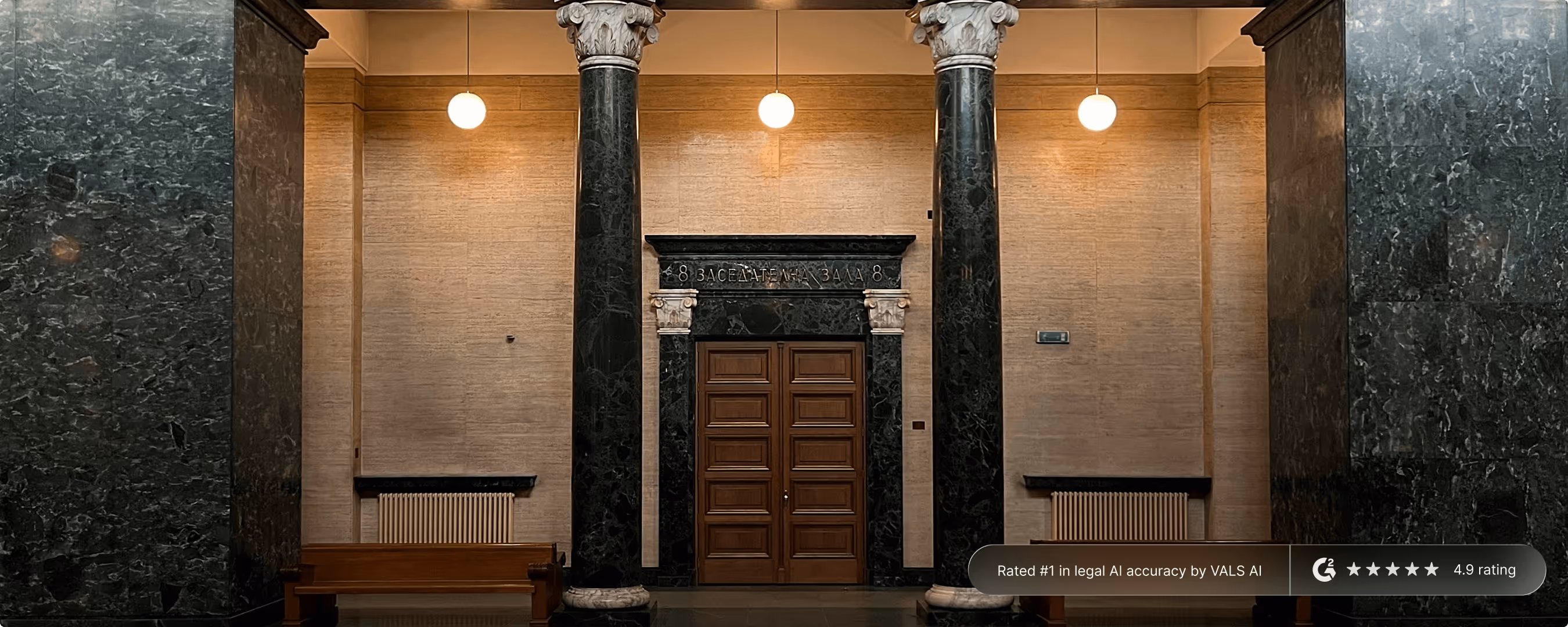 Interior of a courtroom hallway with wooden double doors framed by dark marble columns and beige walls, illuminated by hanging globe lights.