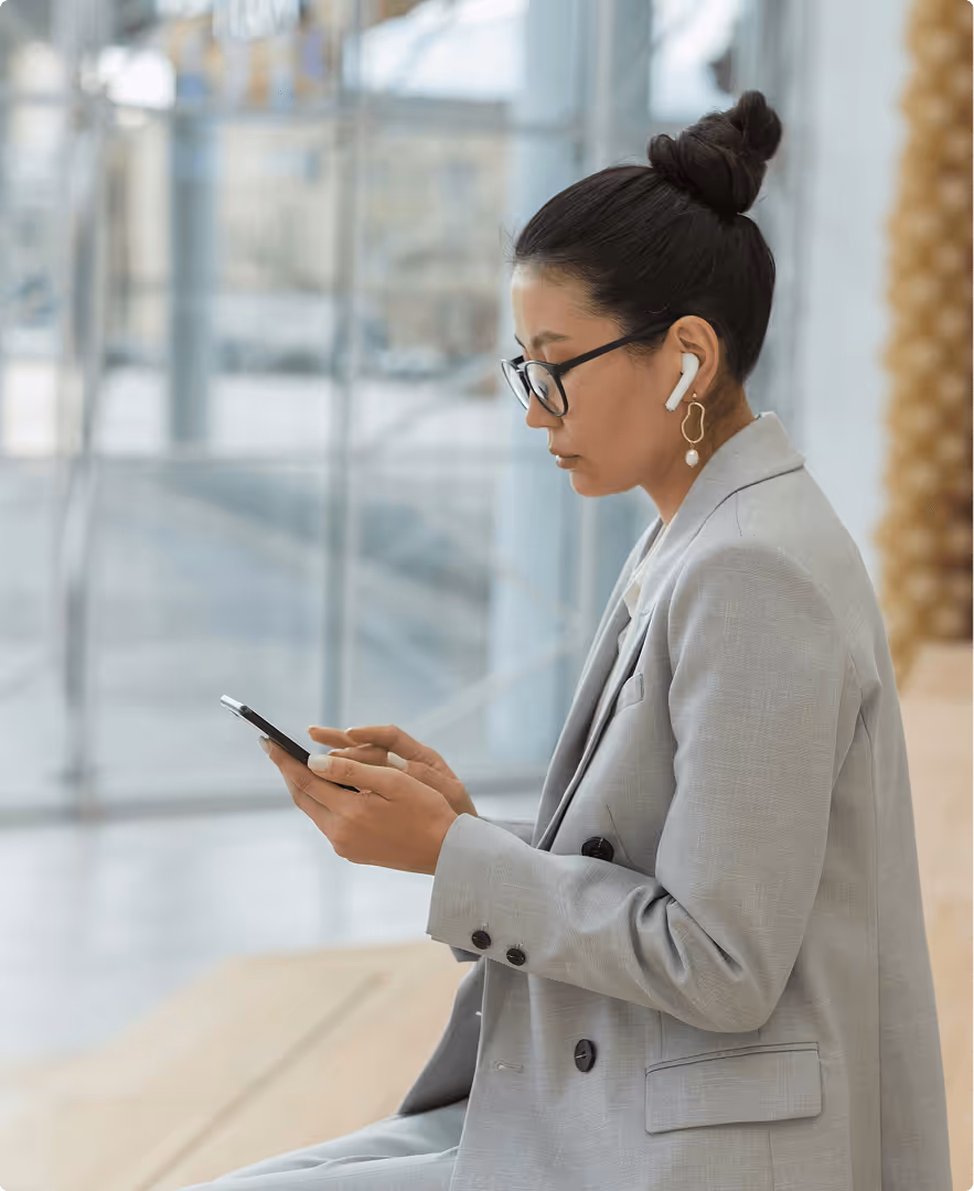 Woman in a gray suit with glasses and wireless earbuds looking at her smartphone indoors.