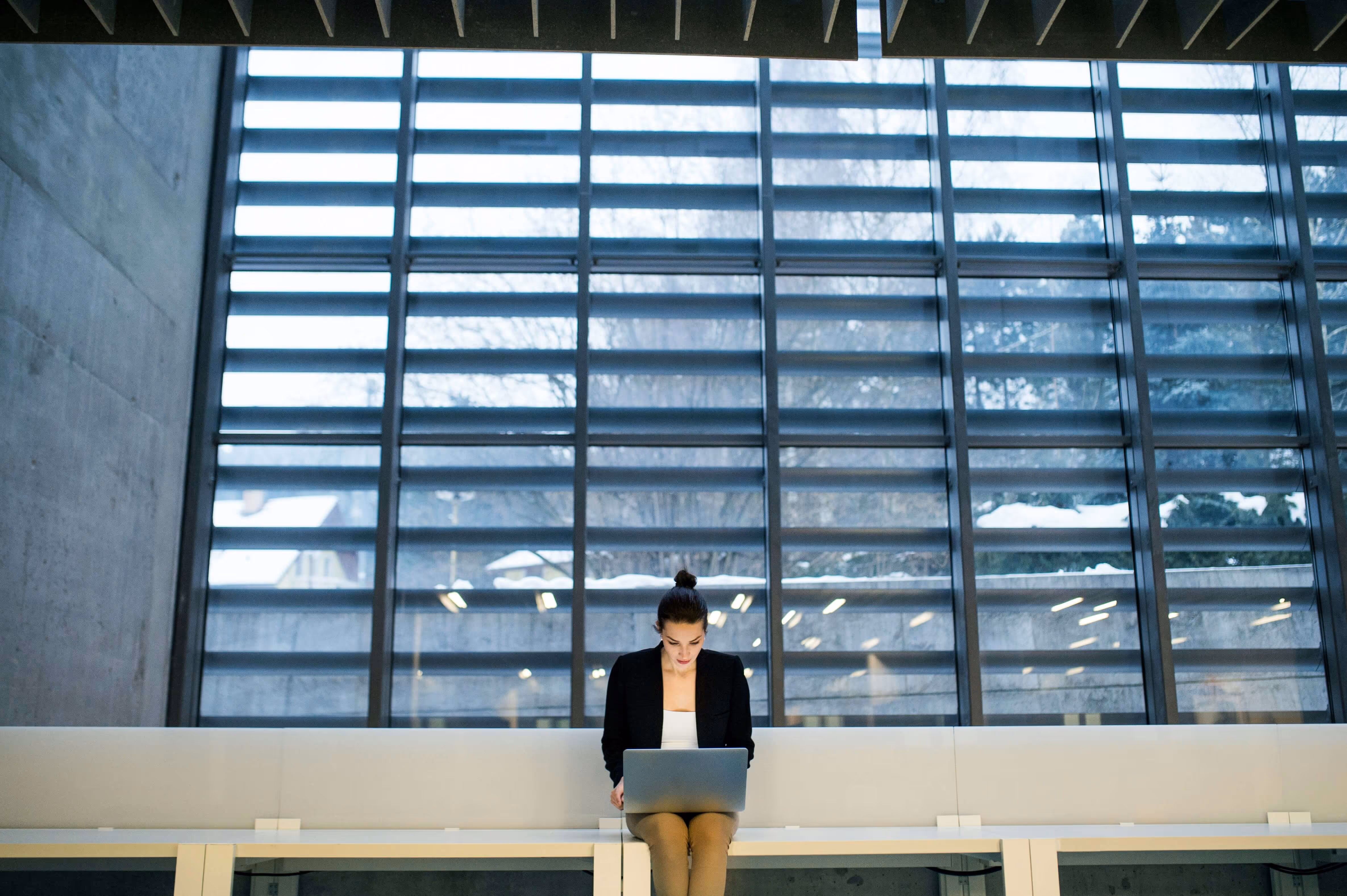 A woman sitting on a long white bench using a laptop in front of large industrial-style windows.
