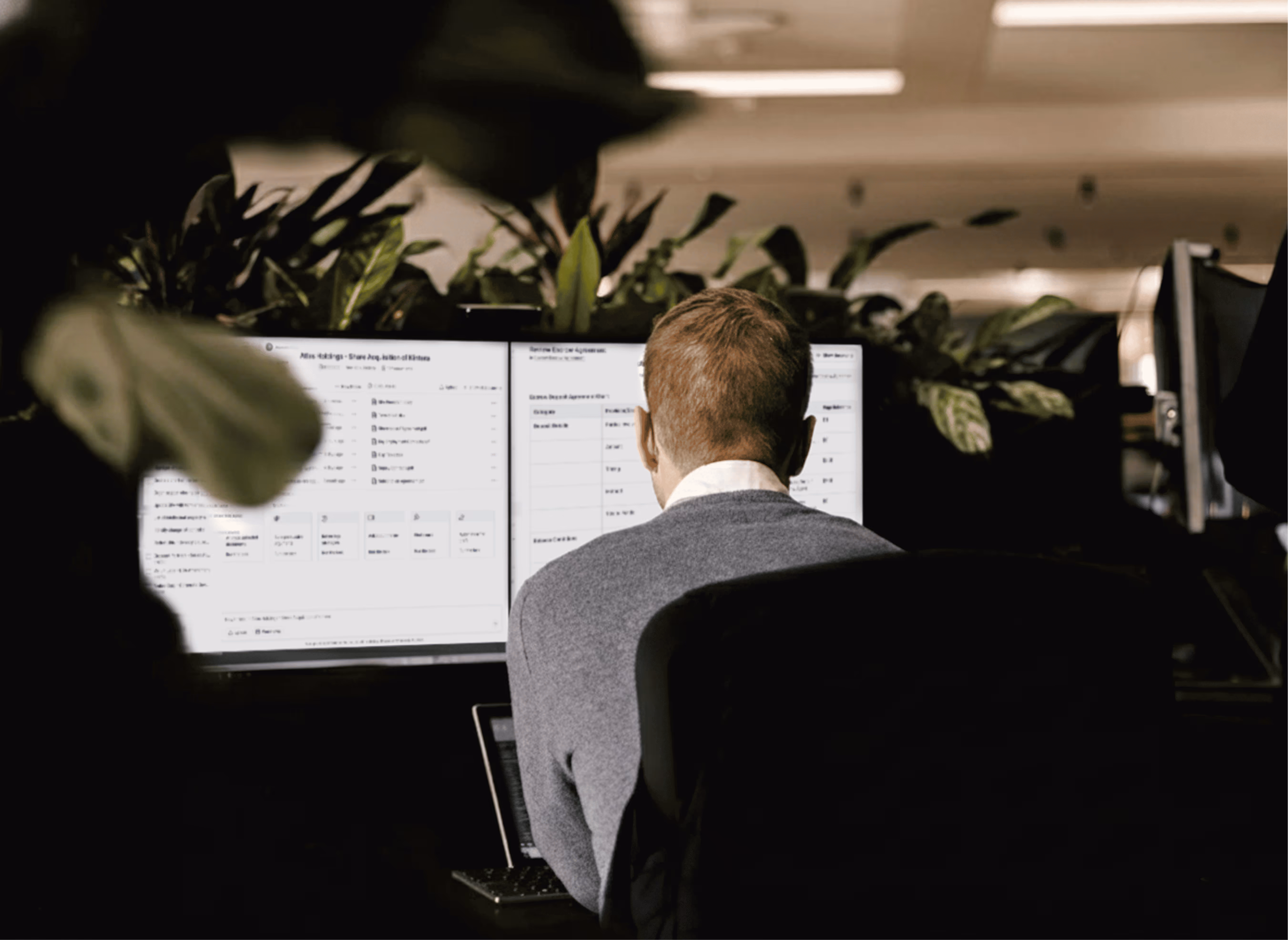 Person with short hair wearing a gray sweater working at a desk with two computer monitors surrounded by plants.