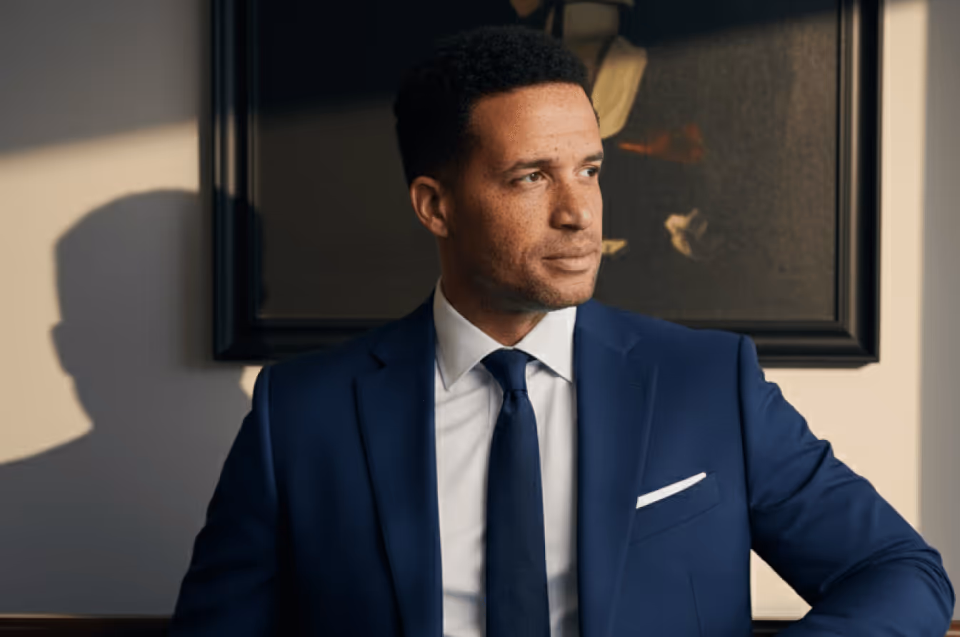 Man in a blue suit and tie looking thoughtfully to the side indoors.