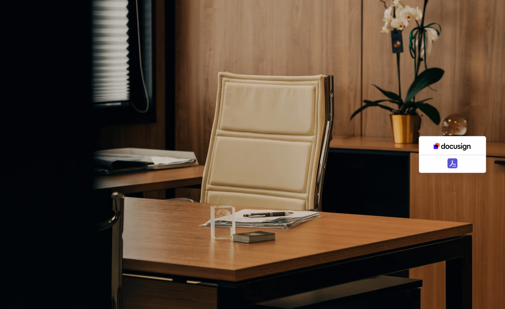 Modern office desk with a beige leather chair, papers and a pen on the desk, and a potted orchid in the background.
