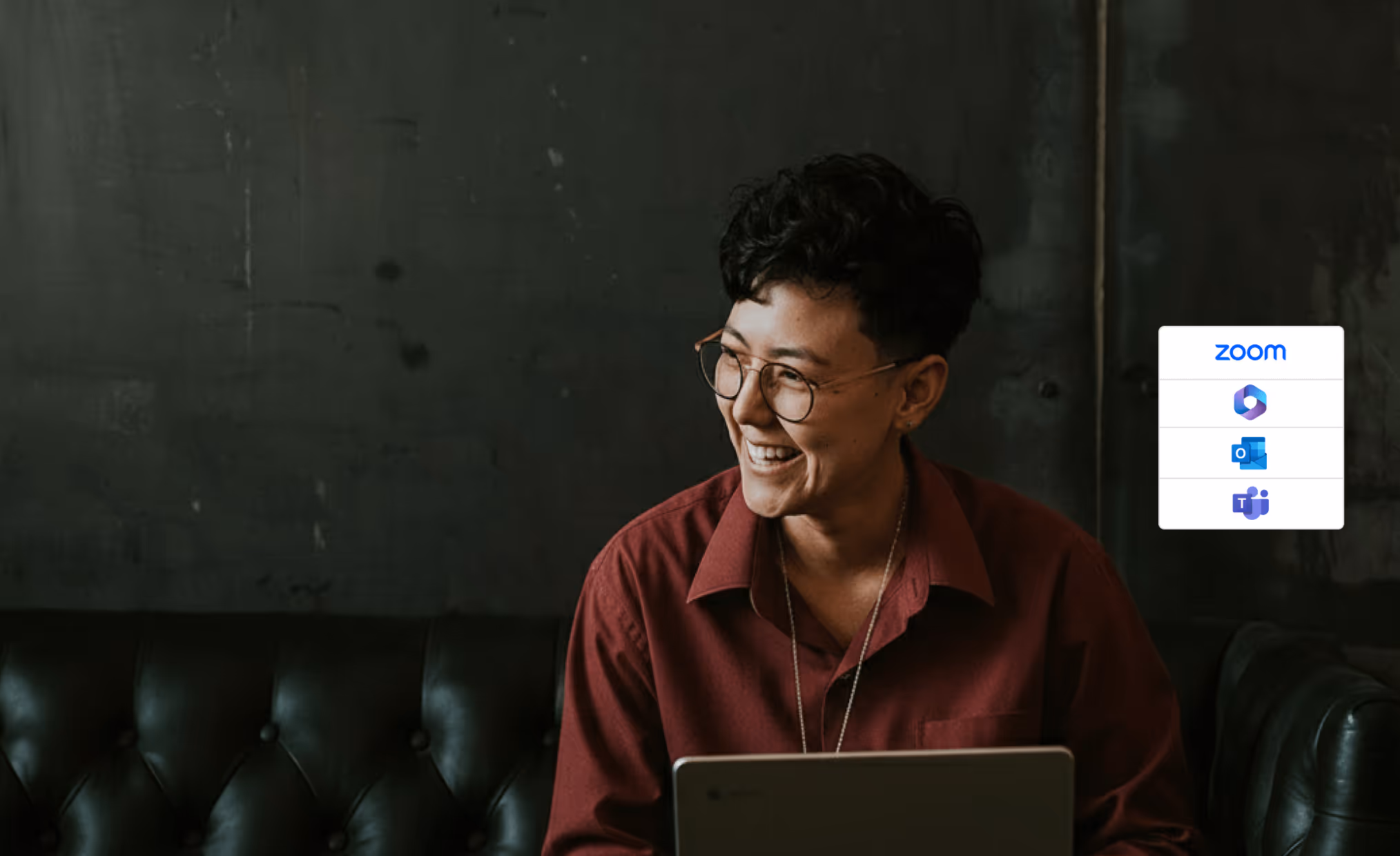 Smiling person with short dark hair and glasses sitting on a black leather couch, working on a laptop with video conferencing app icons overlayed.
