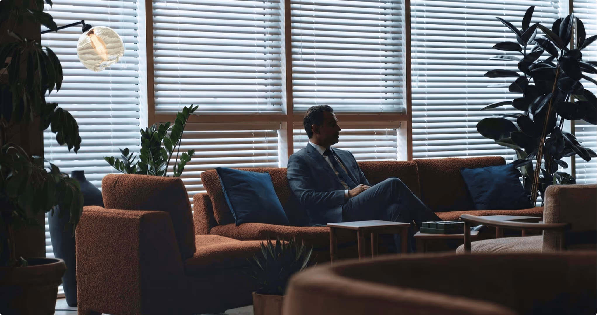 Man in a suit sitting on a brown couch in a dimly lit room with large windows covered by blinds and indoor plants.