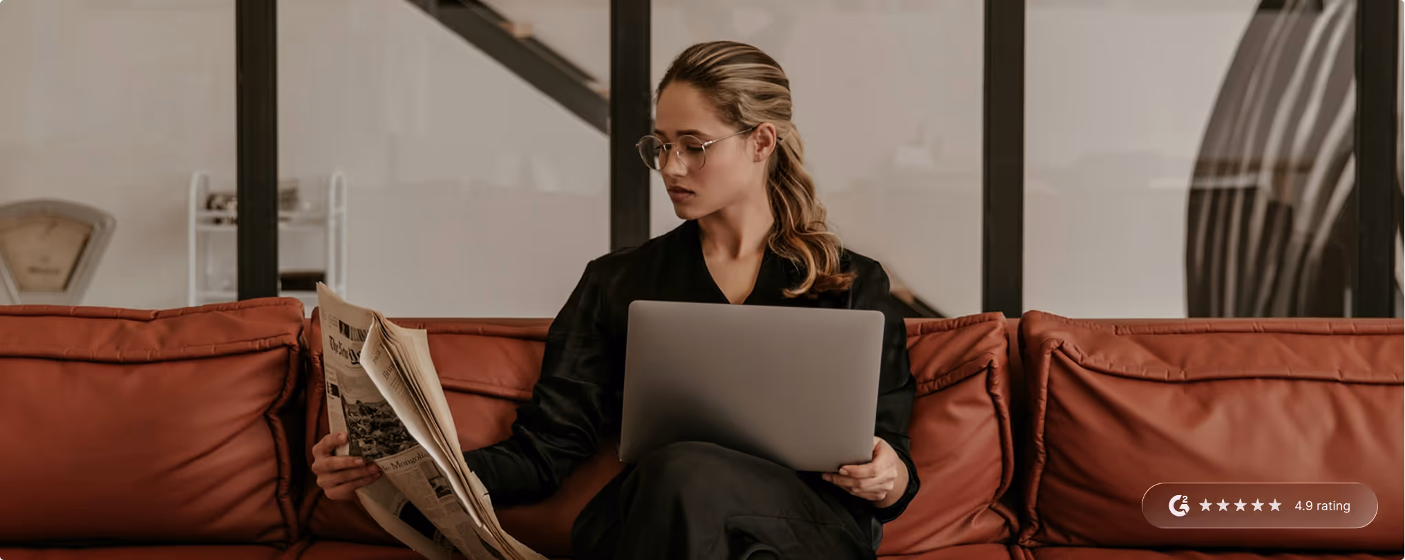 Woman sitting on a brown leather couch reading a newspaper while holding a laptop on her lap.
