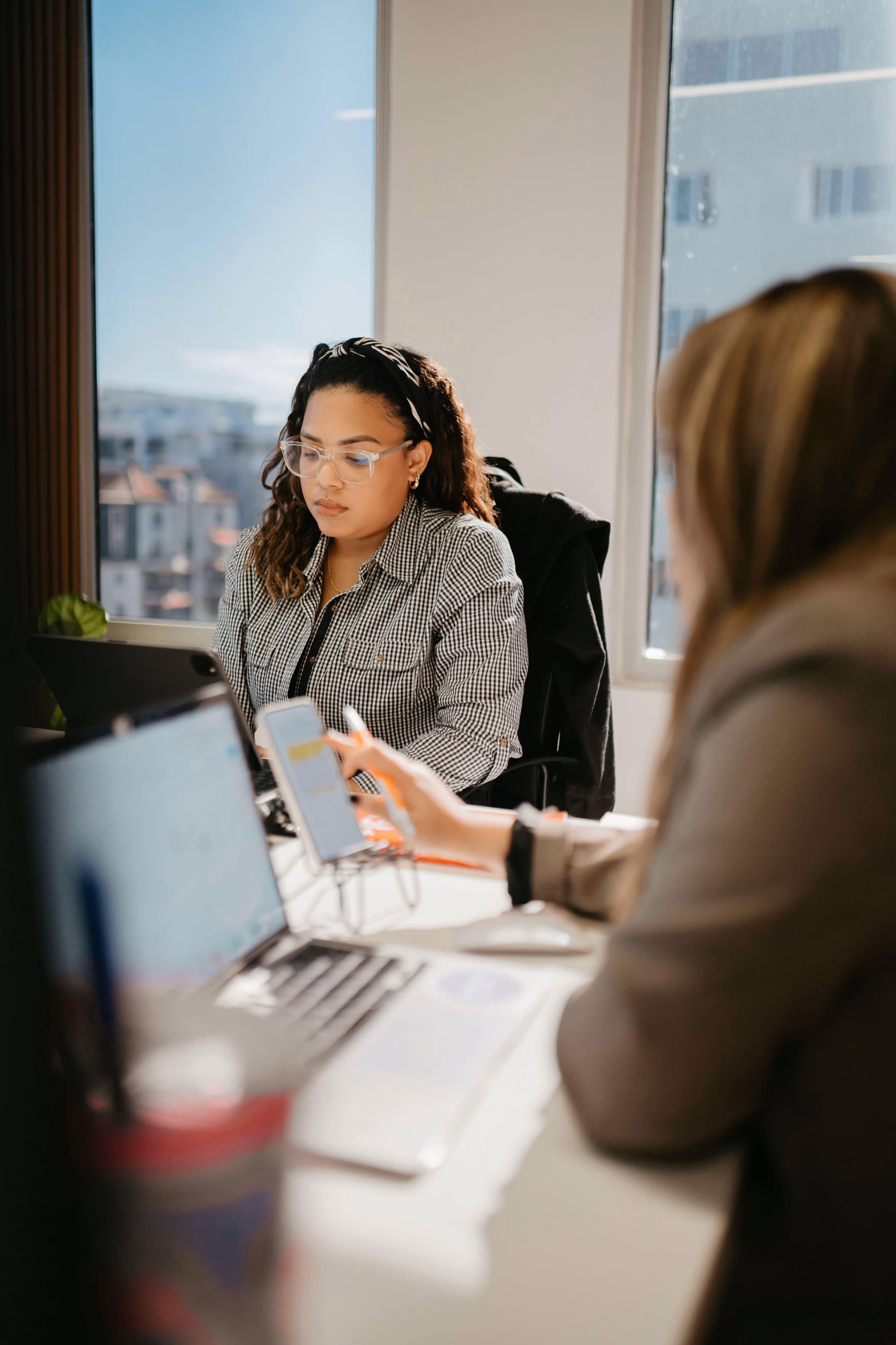 Two women working at a desk with laptops and a smartphone in a sunlit office.