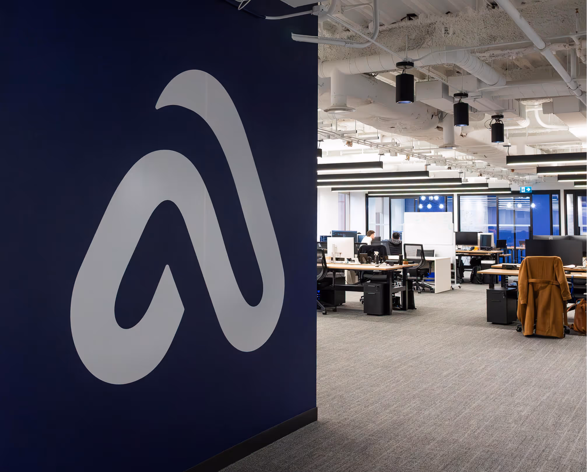 Modern open office space with desks, computer monitors, office chairs, and a large stylized logo on a dark blue wall.