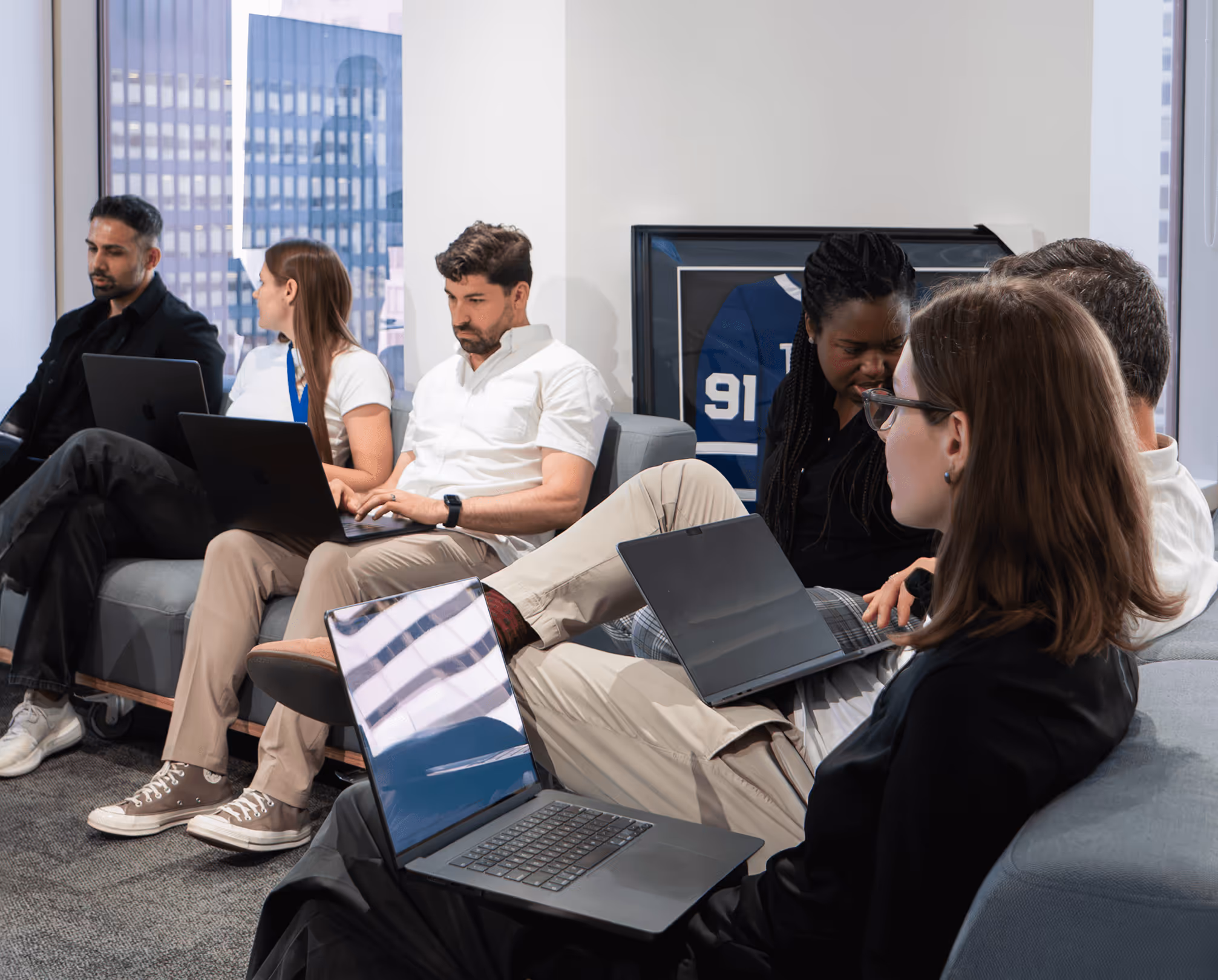 A group of six diverse young professionals sitting on sofas, working on laptops and collaborating in a modern office.