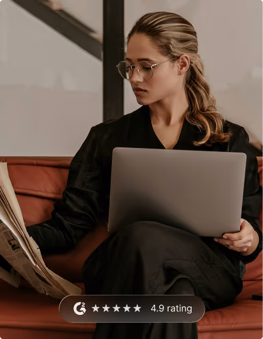 Woman sitting on a brown leather couch reading a newspaper while holding a laptop on her lap.
